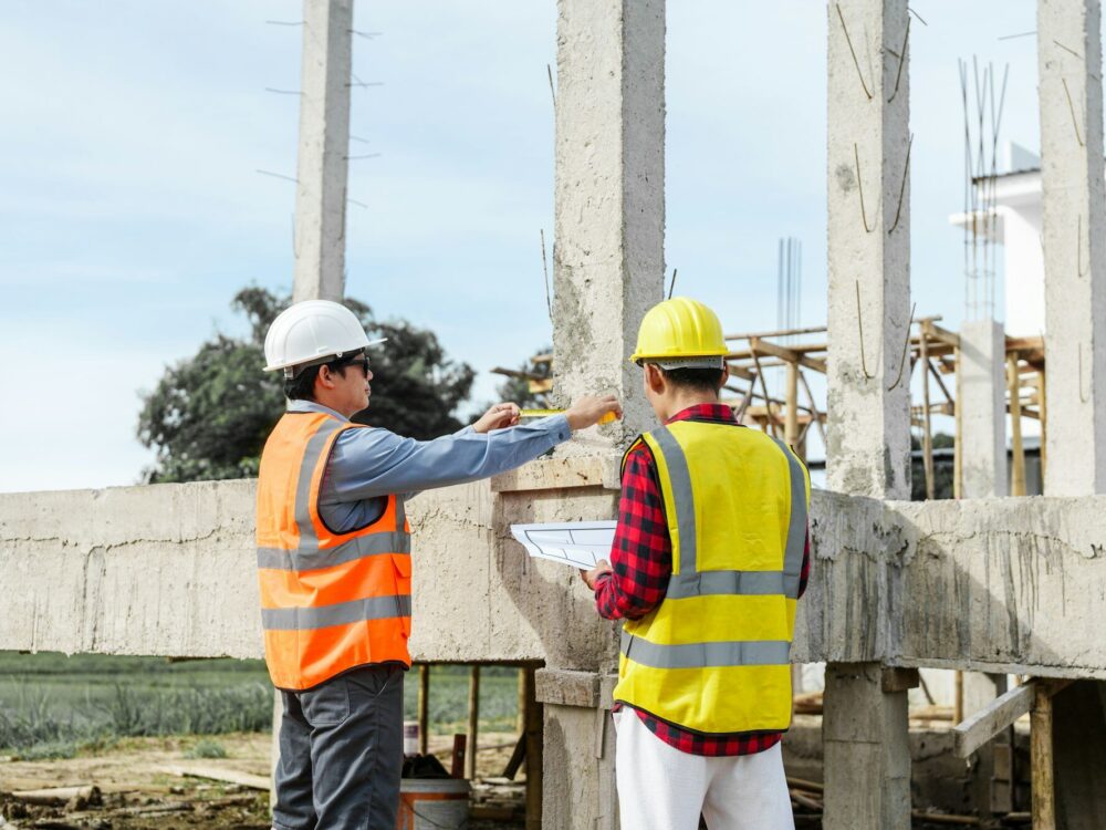 Two male construction workers, including an Asian engineer, meticulously review structural plans